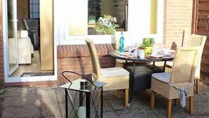 Un patio al aire libre con una mesa, sillas, un vidrio que da a una habitación y un muro de ladrillo.