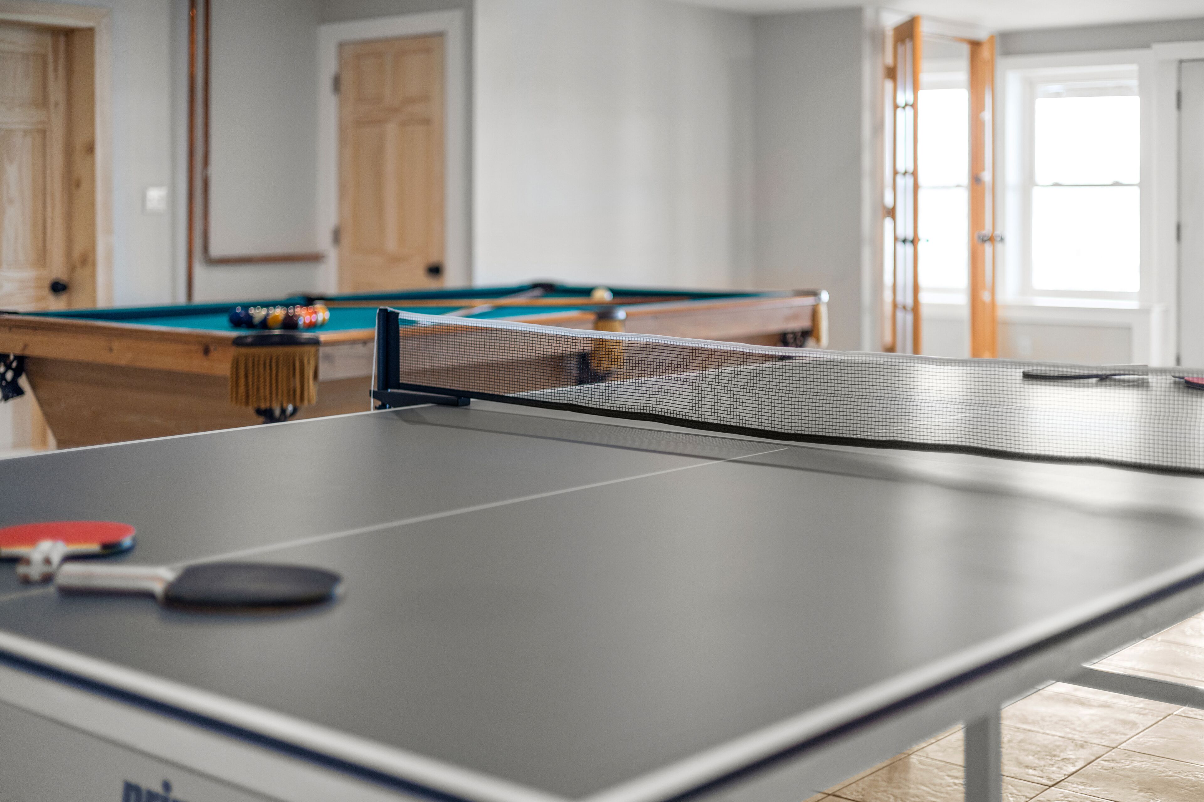 Ein Tischtennis-Tisch mit Schlägern und Bällen in einem Raum mit Holztüren und einem Billardtisch im Hintergrund.