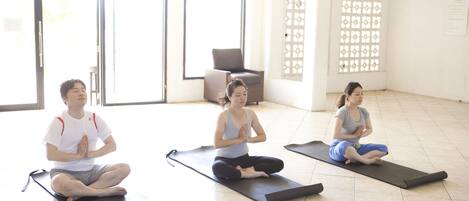 Trois personnes sont assises sur des tapis de yoga dans une pièce dotée de grandes fenêtres et d’une porte.