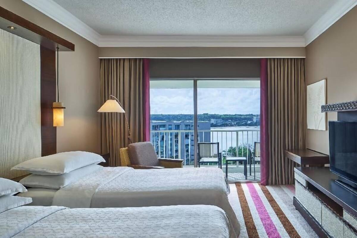 A hotel room with two beds, a TV, a balcony with a view, and a lamp.