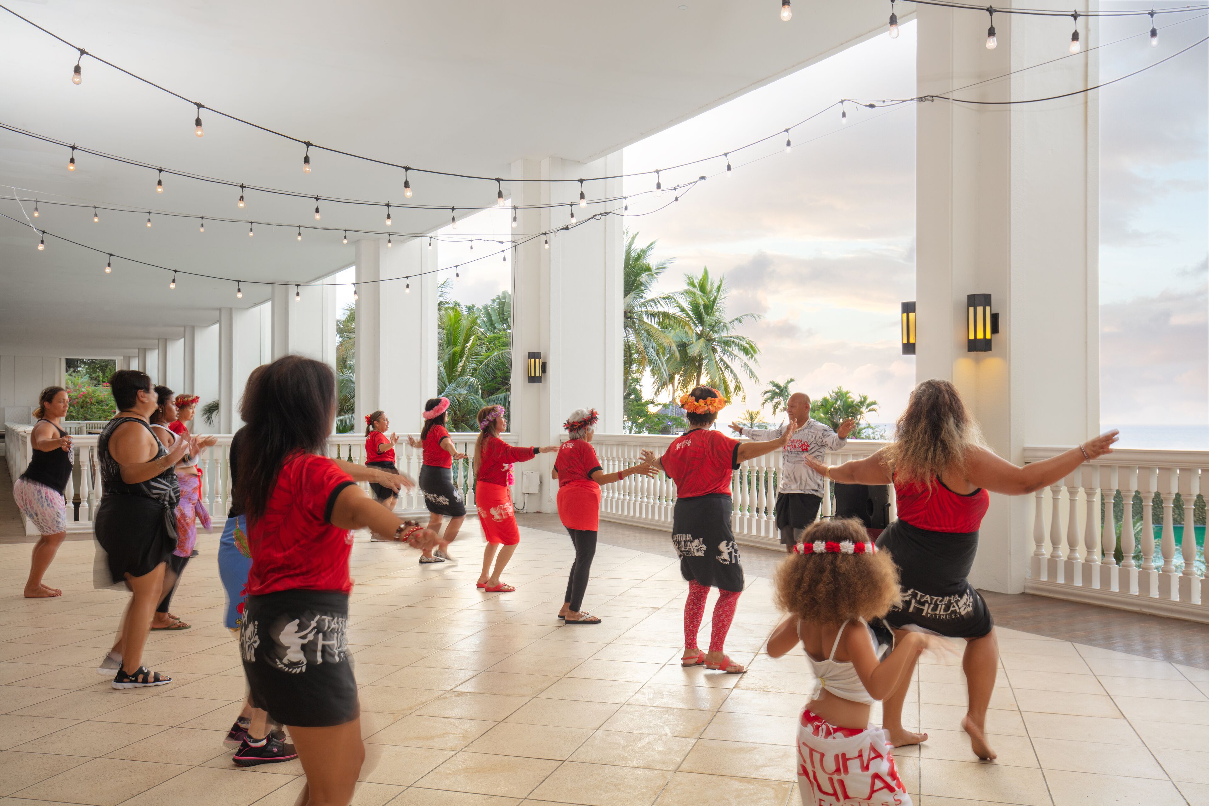 A spacious indoor area with a high ceiling, string lights, and a balcony overlooking palm trees.