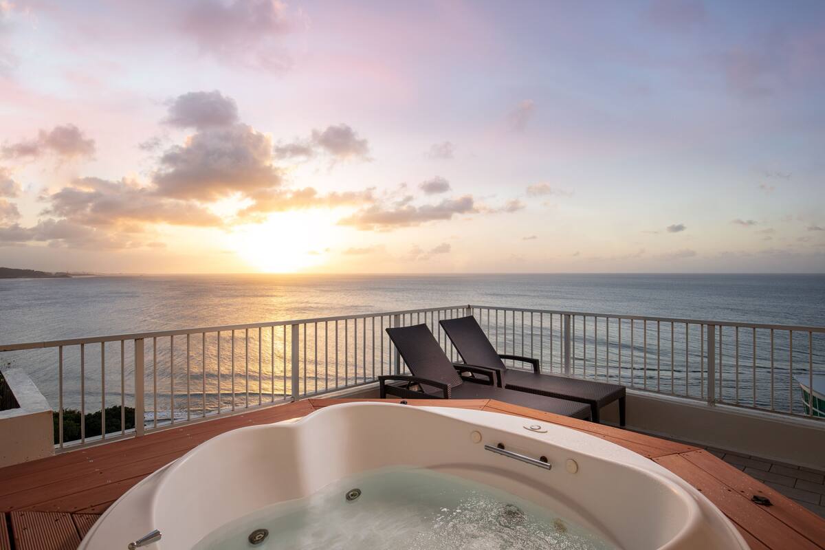 A balcony with a hot tub, lounge chairs, and a view of the ocean at sunset.