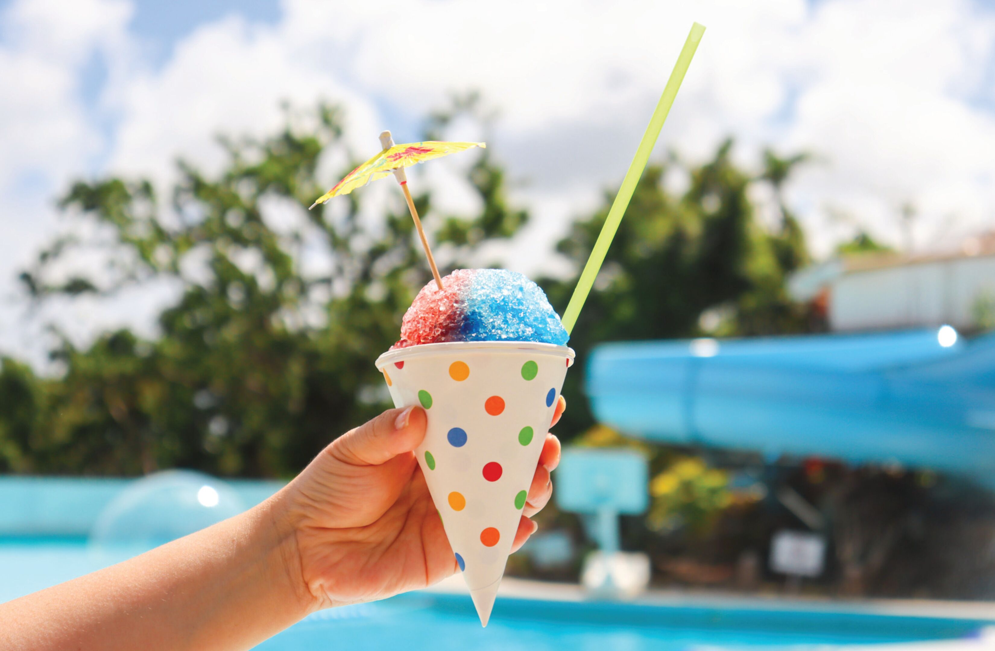 Une main tenant une glace à la vanille à pois, avec une paille rayée et un petit parasol, le tout sur fond de piscine.