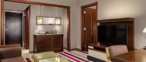 A hotel room with a sofa, a glass-top coffee table, a television, a minibar, and a wooden cabinet.