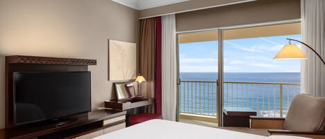 Habitación de hotel con una cama grande, un televisor de pantalla plana, un escritorio con lámpara y vistas al océano a través de una puerta corredera.