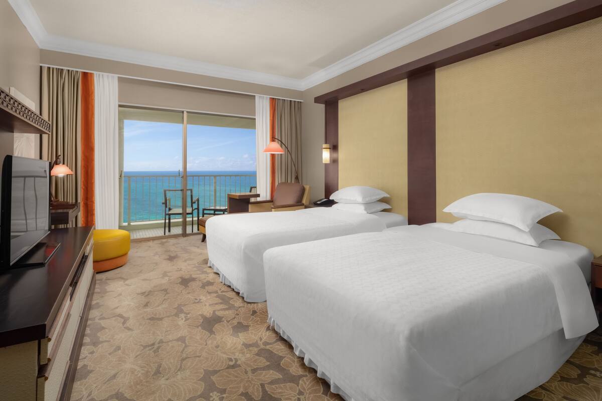 A hotel room with two beds, a TV, a desk, and a balcony with a view of the ocean.