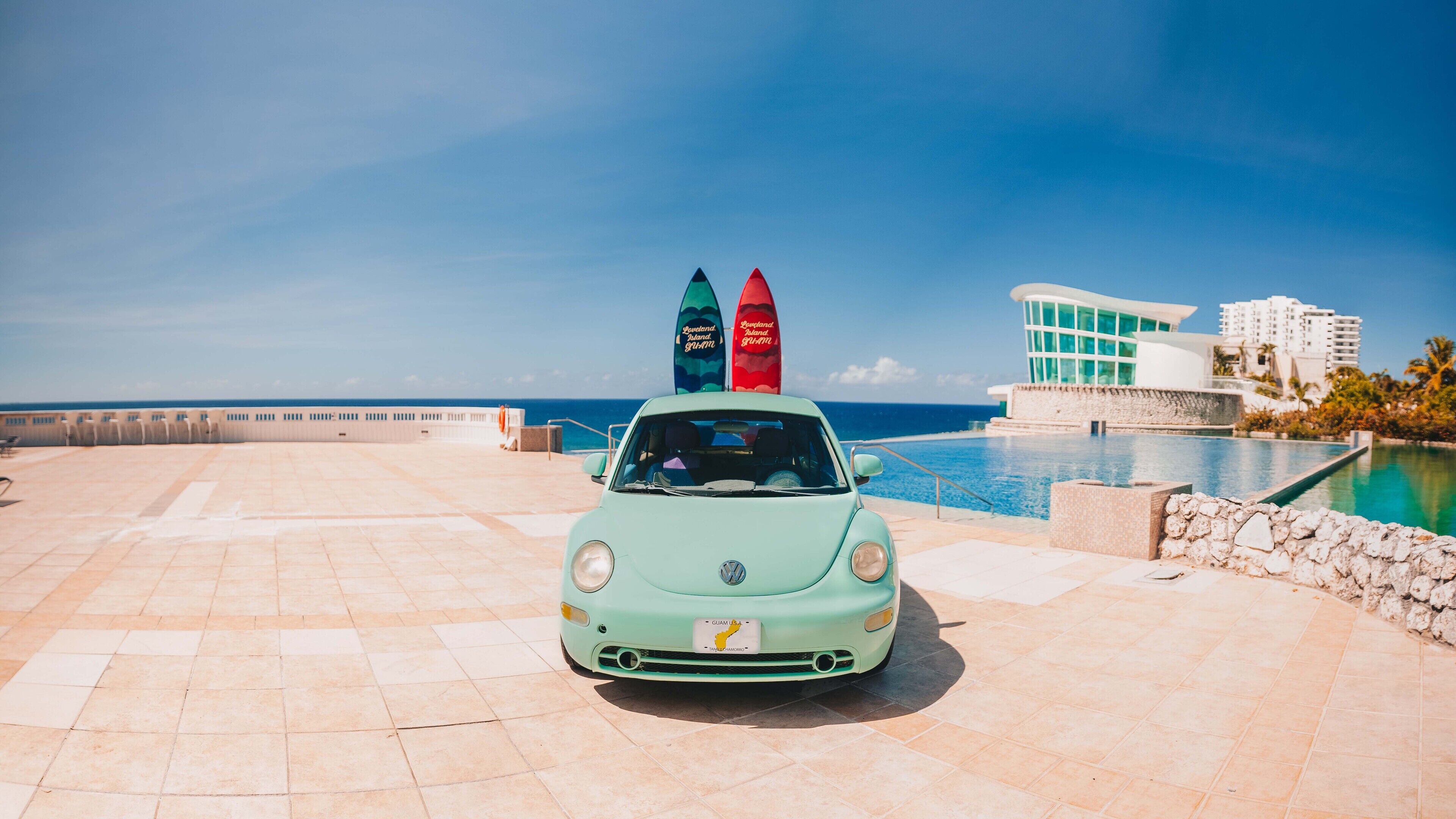 Un coche Volkswagen clásico estacionado en una terraza con baldosas, una piscina y vista al océano.