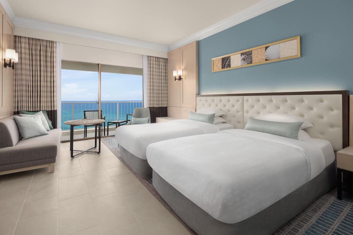 A hotel room with two beds, a sofa, a small table, and a view of the ocean.