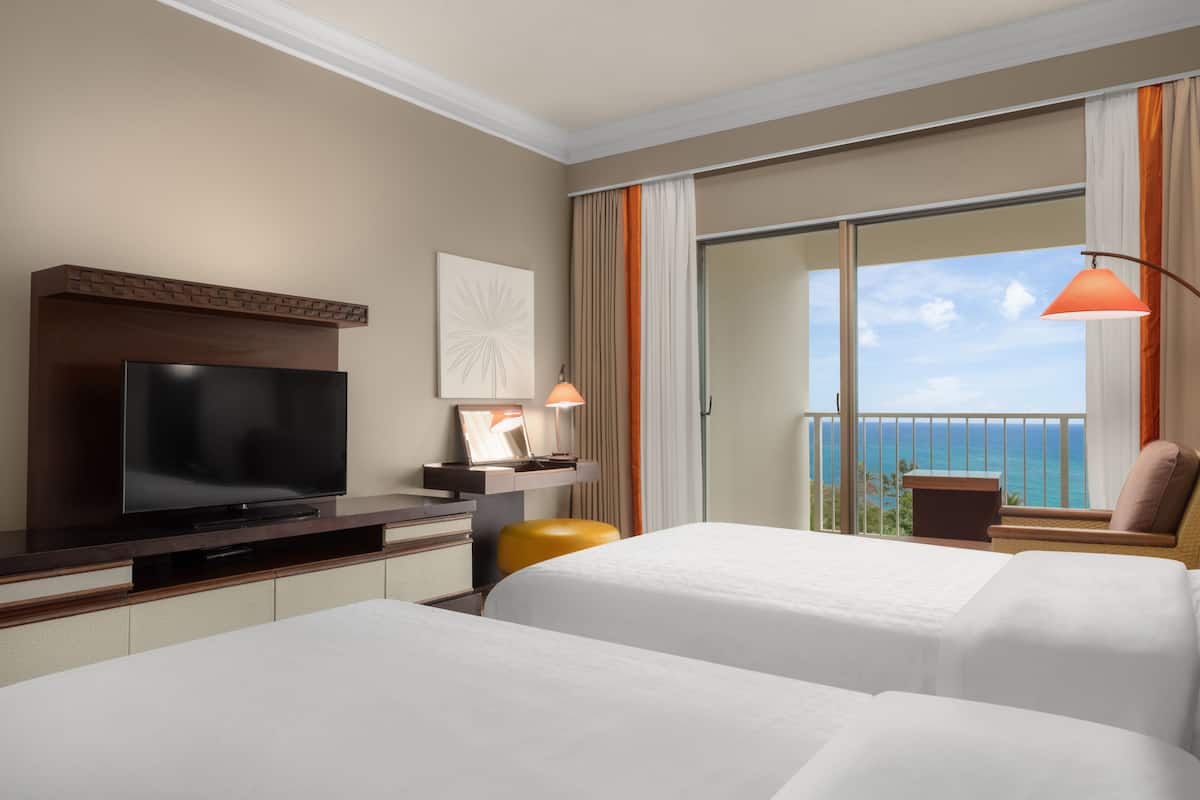 A hotel room with a flat-screen TV, two beds, a desk with a lamp, and a balcony with a view of the ocean.