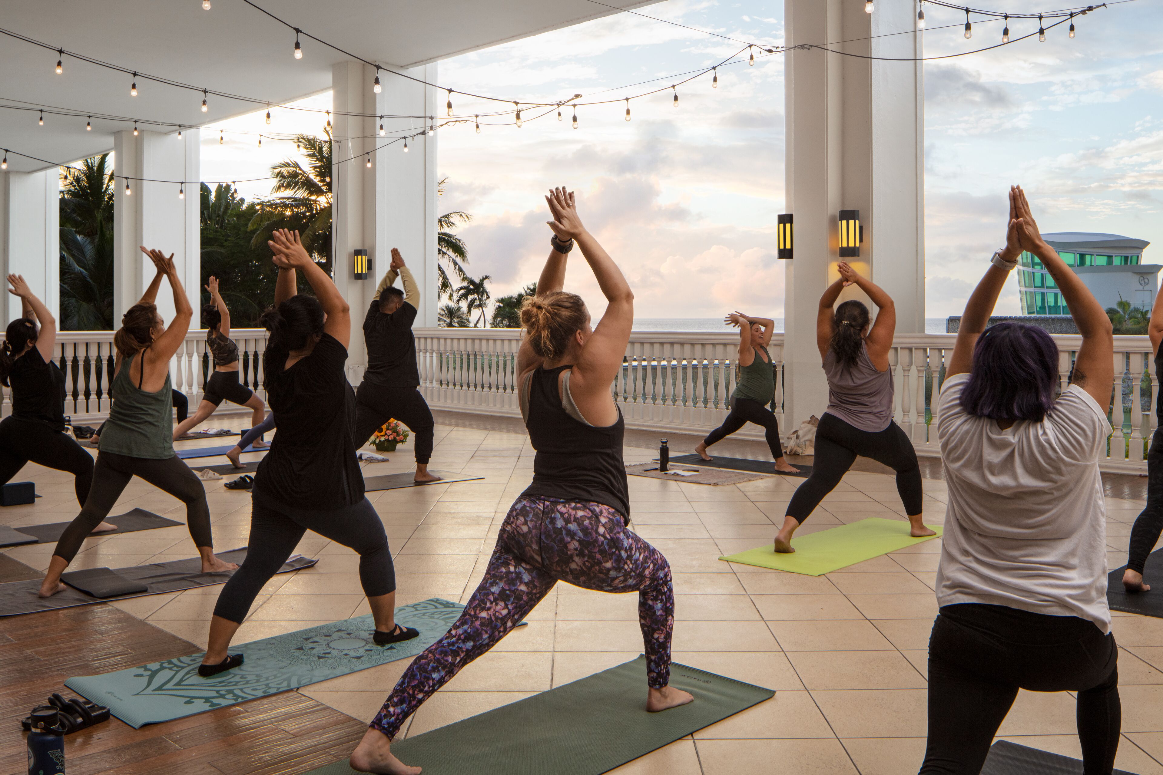 A group of people performing yoga on mats in an outdoor setting with a view of the ocean and palm trees.