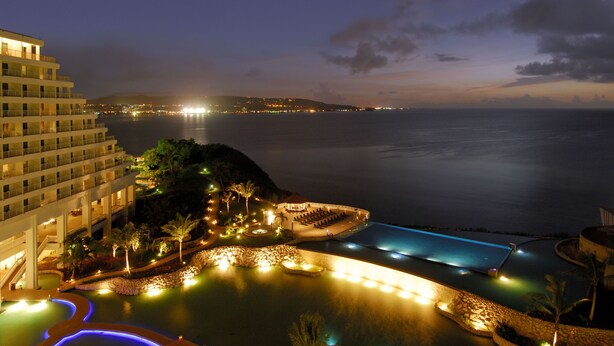 A resort with a pool area, a building with multiple floors, and a view of the sea at night.