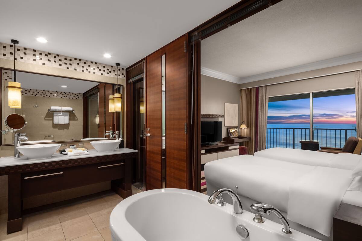 A hotel room with a large bathroom, a bed, a TV, and a view of the ocean.