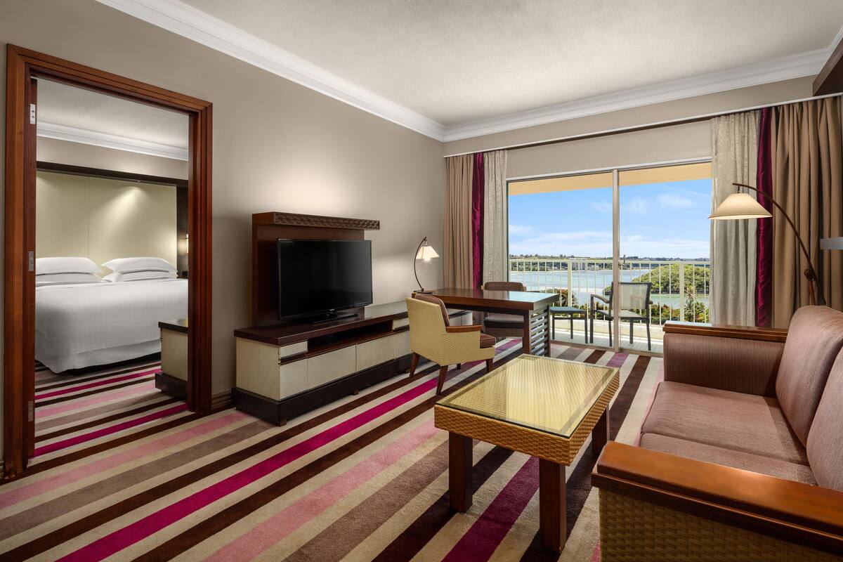 A hotel room with a bed, a TV, a sofa, a dining table, and a view of the sea.