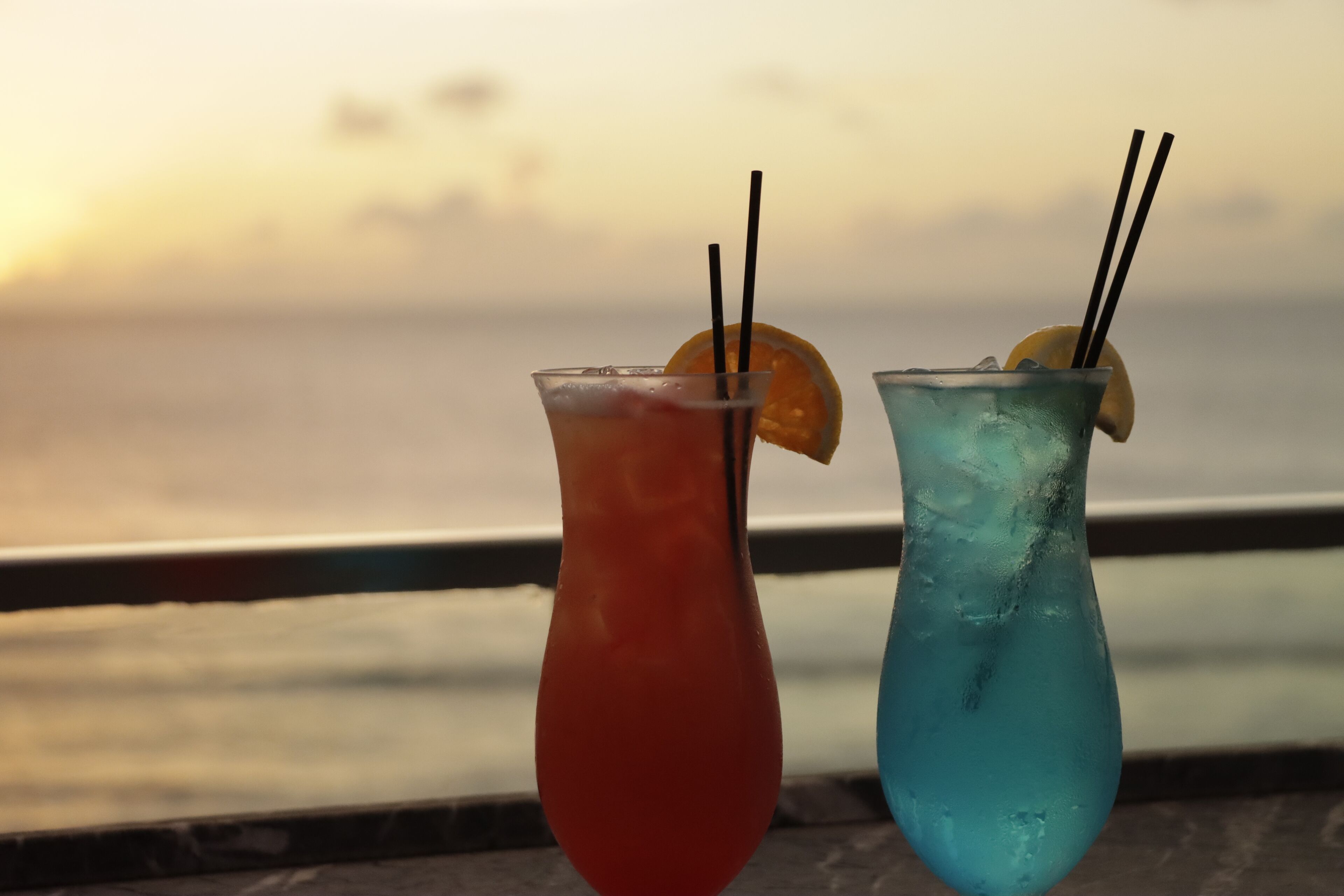 Two cocktails with straws and lemon slices, set against a sunset view.