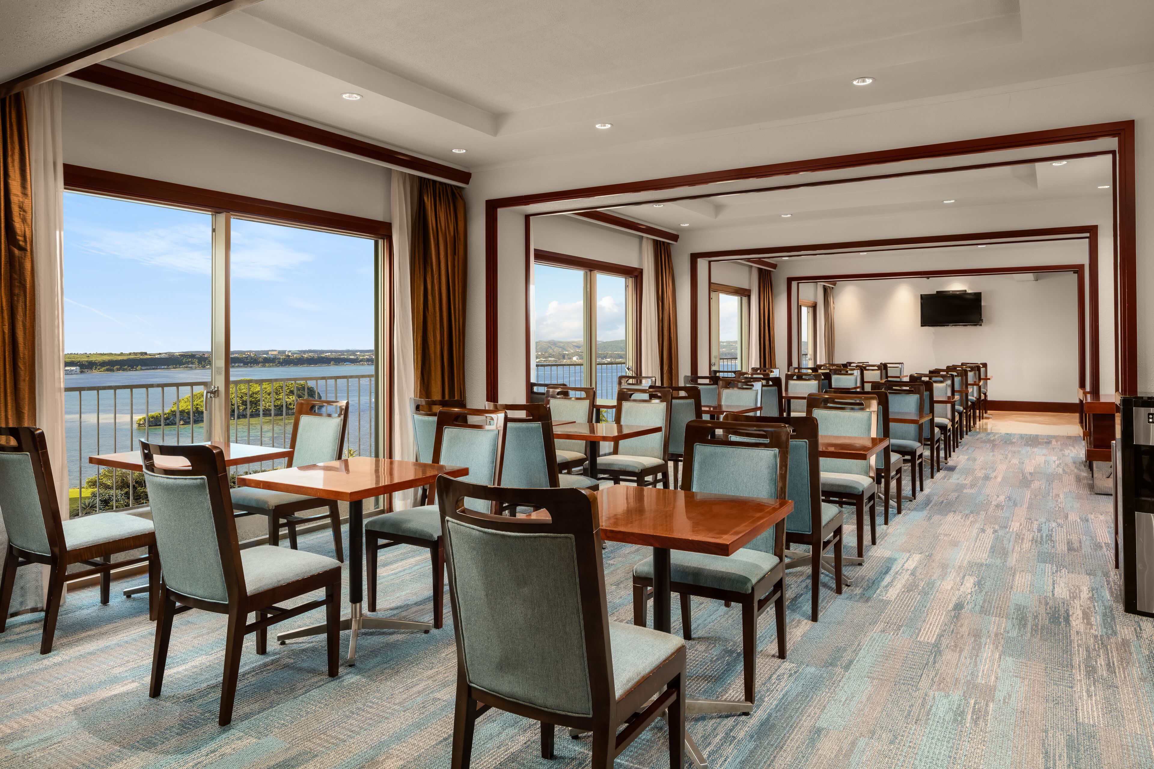 A hotel dining area with tables and chairs, large windows with curtains, and a view of the water.