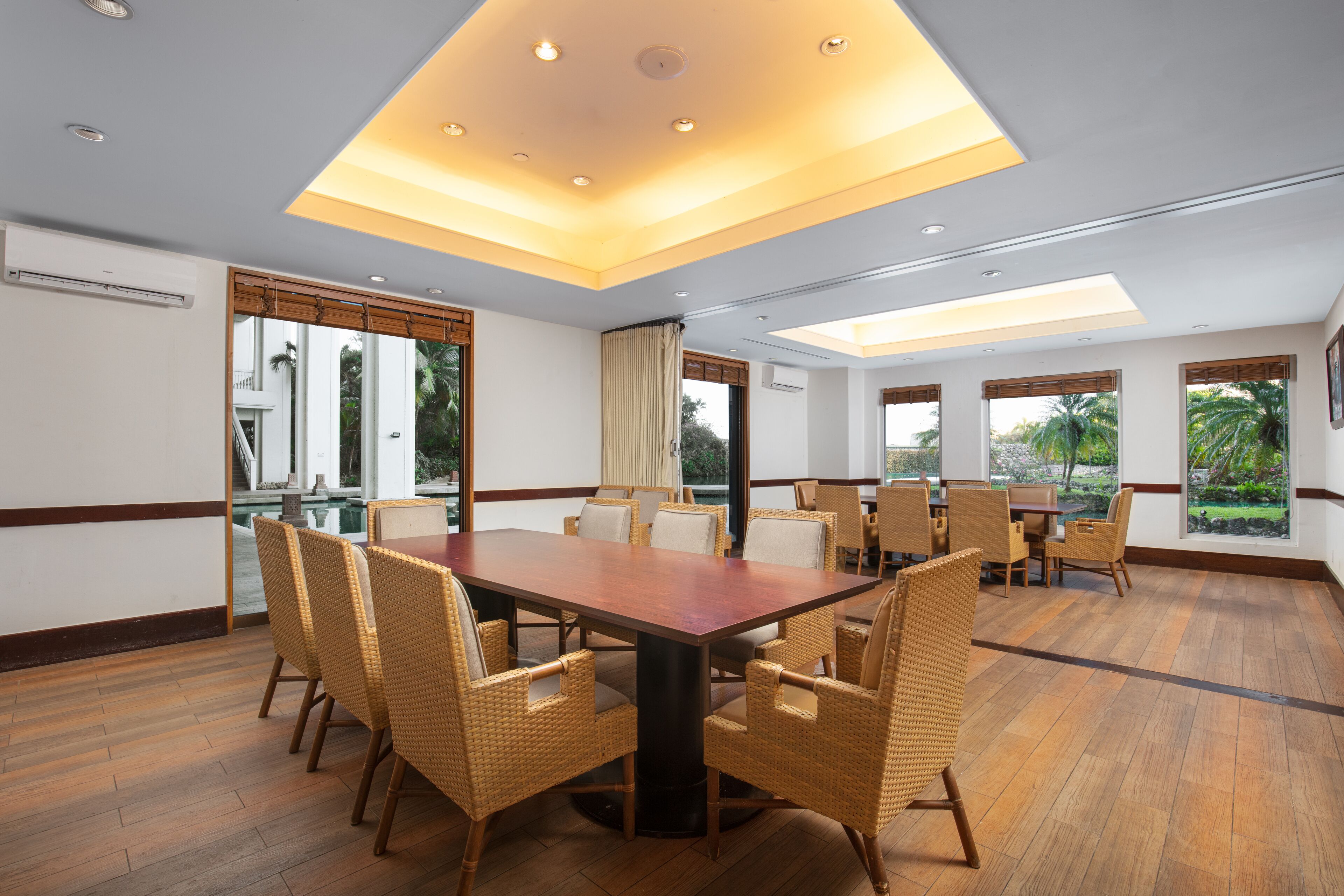 A conference room with a large wooden table and wicker chairs, large windows with views of greenery, and recessed ceiling lighting.