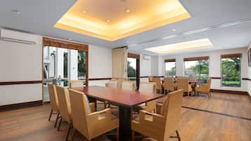 Un salón de conferencias con una amplia mesa de madera y sillas de mimbre, ventanas grandes con vistas a la vegetación e iluminación empotrada en el techo.