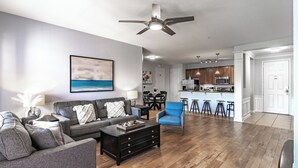 A modern living room with a grey sofa, a blue armchair, and a coffee table. There is a ceiling fan, a painting of a beach, and a kitchen area with bar stools. - ★SPACIOUS 4BD/2BA★CONVENTION CENTER/ UNIVERSAL EPIC UNIVERSE WALKING DISTANCE (Orlando)