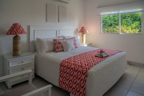A bedroom with a white bed, red patterned pillows, a white nightstand, and a window with a view of greenery.
