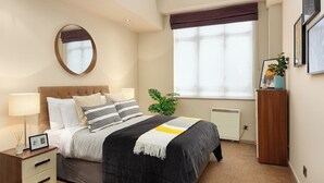 A bedroom with a bed, bedside tables, a mirror, a window with blinds, a radiator, and a small plant. - Lawrence House by Interhome (London City)