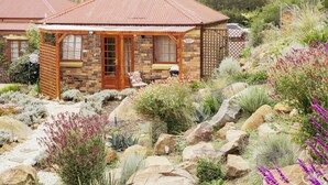 Una cabaña rústica con fachada de piedra, porche de madera y un pequeño jardín con diversas plantas y rocas.