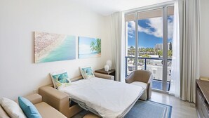 A hotel room with a bed, a sofa, a chair, and a large window with a view of palm trees and buildings. - CH Luxury Spacious Beach Condo | Ocean Breeze-CS40 (Hollywood)