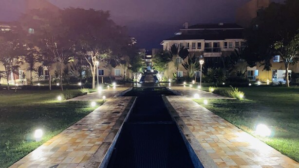 A nighttime view of a well-lit pathway leading to a building with a pool, surrounded by trees and illuminated by street lamps. - The Sun (2 bedrooms in the heart of Brasilia) (DF)