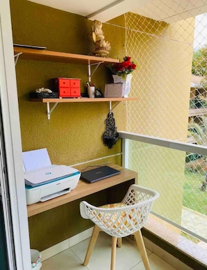Varanda com escrivaninha, cadeira, prateleira pequena com objetos decorativos e vista para vegetação.