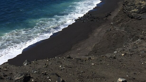 A coastal landscape with a curved black sand beach, ocean waves, and a clear blue sky. - Fun Family Private Retreat with lighted Sports Court and Pool (Ocean View)