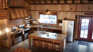 A wooden cabin-style kitchen with a central island, bar stools, a refrigerator, and a window overlooking a snowy landscape. - Fun Family Private Retreat with lighted Sports Court and Pool (Ocean View)