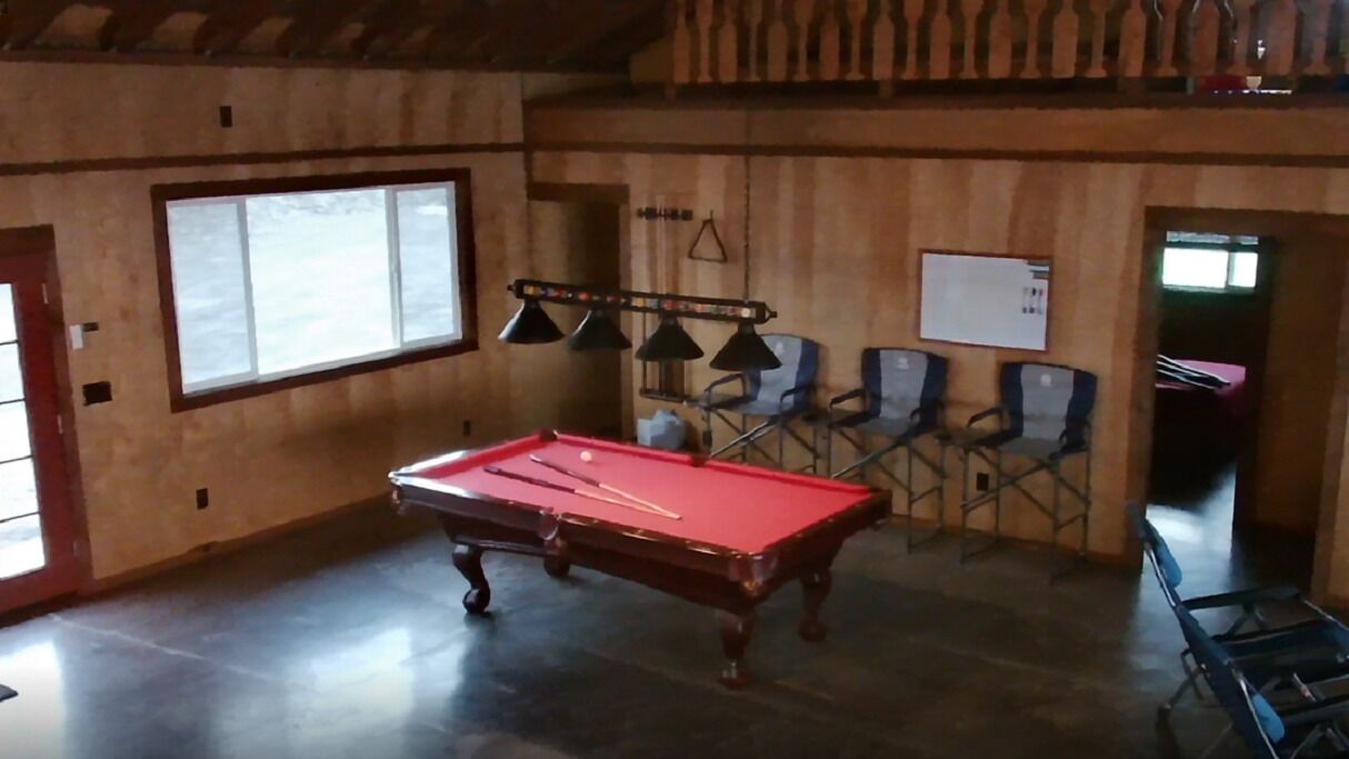Une table de billard dans une pièce aux murs en bois, avec une grande fenêtre et plusieurs chaises.