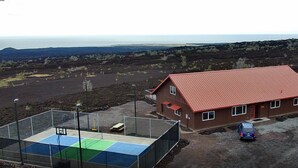 A house with a red roof and a fenced tennis court in front. - Fun Family Private Retreat with lighted Sports Court and Pool (Ocean View)
