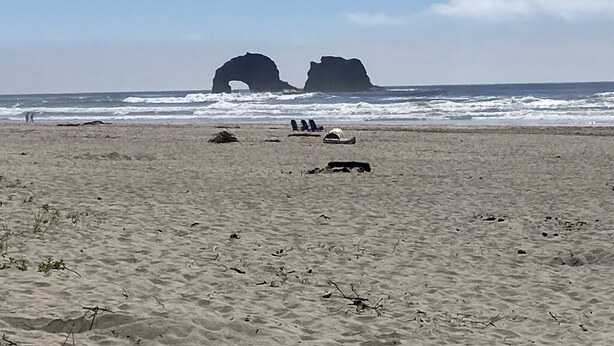 A sandy beach with two rock formations in the distance and a few people and sun loungers in the foreground.