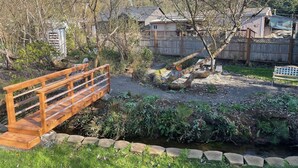 A wooden bridge over a stream in a garden with a wooden fence and houses in the background.