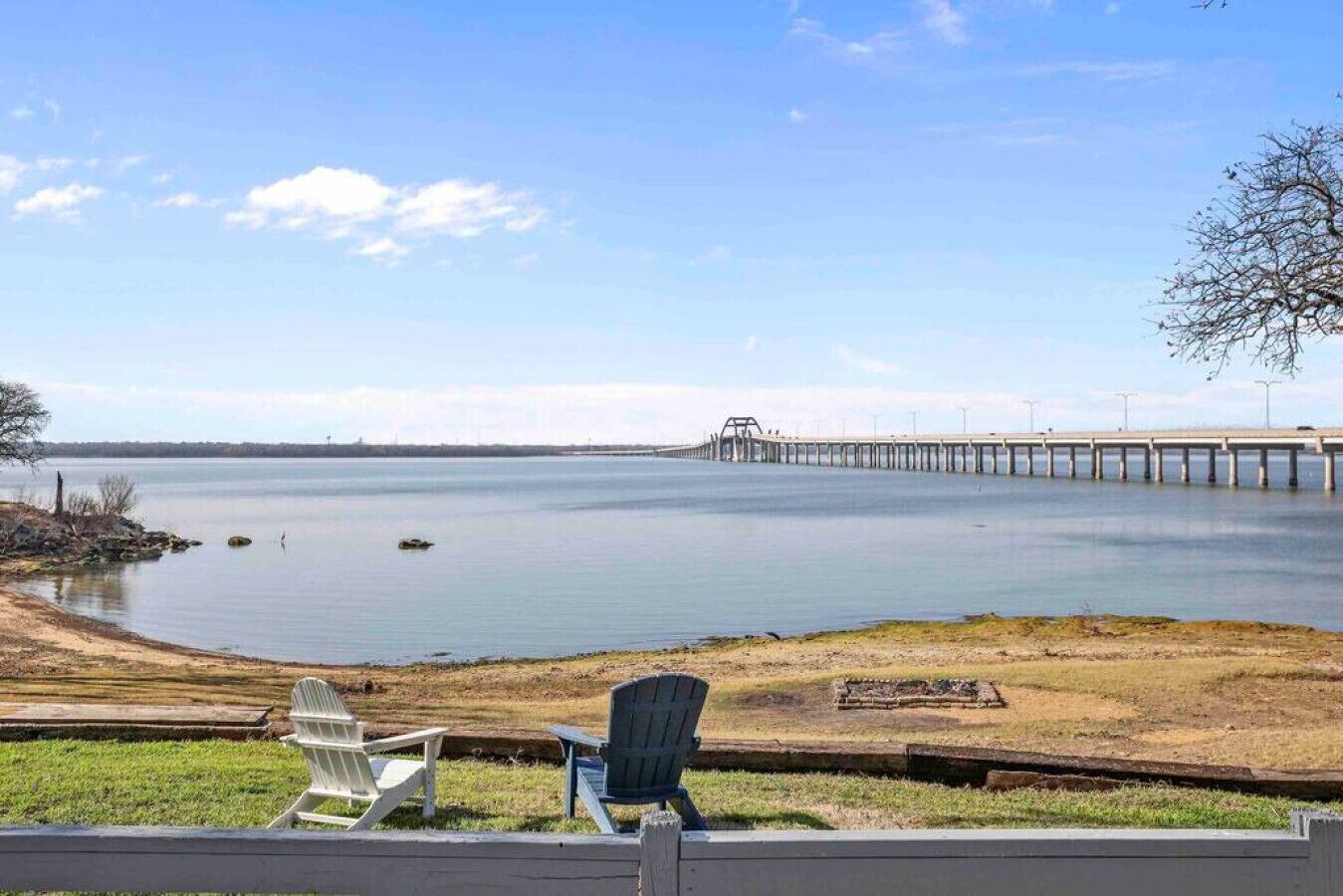 A lakeside view with a pier extending into the water, two Adirondack chairs, and a grassy area in the foreground.