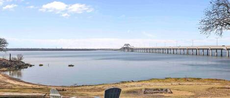 A lakeside view with a pier extending into the water, two Adirondack chairs, and a grassy area in the foreground.