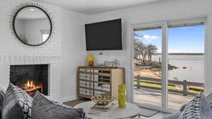 A living room with a fireplace, a flat-screen TV, a large window with a view of a lake, and a cozy seating area. - Rare Modern Beachfront Lake House on Lewisville Lake with Hot Tub, Deck & BEACH (Little Elm)