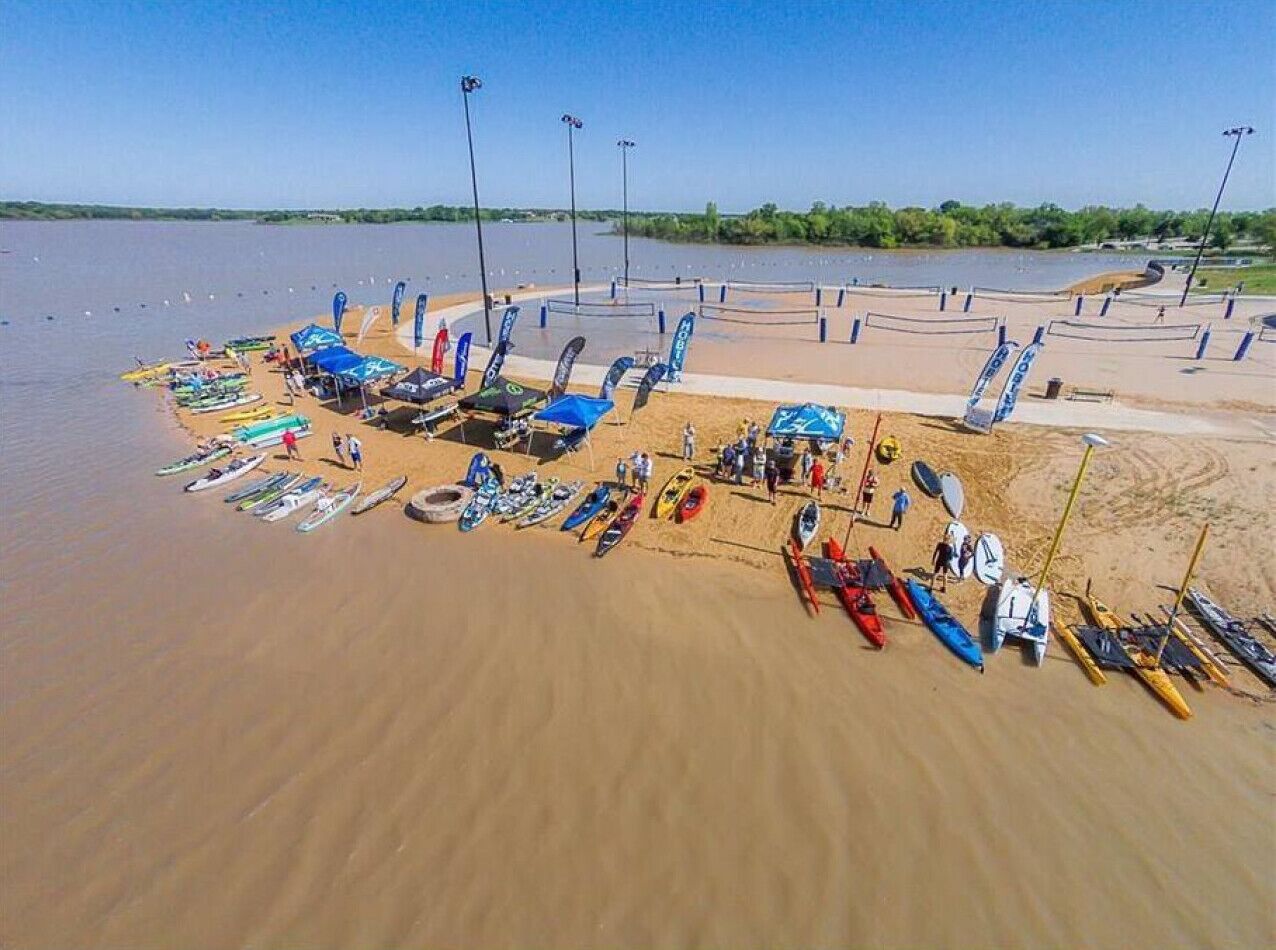 Luchtbeeld van een strand met vele kajaks en kano's op de zandstrook, waarbij enkele mensen in de buurt staan.