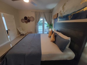 A bedroom with a bed, a chair, a ceiling fan, a window with curtains, and a wall with a painting. - BluWaves ~ Oceanfront Condo minutes from Ocho Rios (Tower Isle)