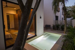 A modern hotel room with a glass door leading to a balcony, a small pool, and a seating area with outdoor furniture.