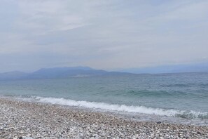 A pebbly beach with calm sea waves and distant mountains under a cloudy sky.