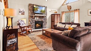 A cozy living room with a stone fireplace, a large flat-screen TV, a ceiling fan, and a comfortable brown sofa. - “THE ONE” Chalet, Perfect Getaway w/ Hot Tub, Multicade, Pool Table (Albrightsville)