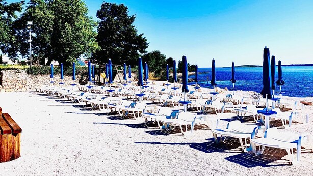 Een strand met rijen witte ligstoelen en blauwe parasols, zand en een heldere blauwe zee.