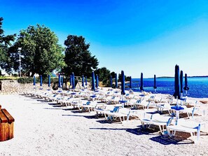 A beach with rows of white lounge chairs and blue umbrellas, a sandy beach, and a clear blue sea.
