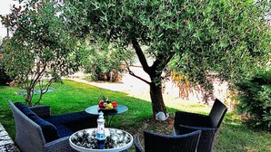 Een terras met rieten meubels en een ronde tafel, omgeven door een verzorgd gazon en bomen.