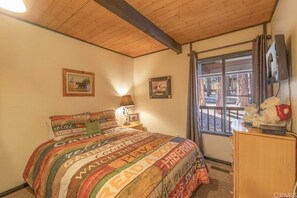 A bedroom with a wooden ceiling, a bed with a colorful striped bedspread, a nightstand with a lamp, a dresser with stuffed animals, and a window with curtains.