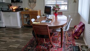 A dining area with a round table, chairs, and a rug. There is a kitchen in the background with cabinets and appliances.