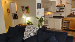 A living room with a dark blue sofa, a small table with a lamp, and a kitchen in the background.