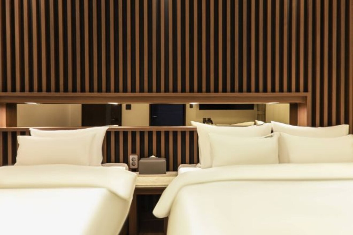 Una habitación de hotel con dos camas, cabecera de madera con listones y mesita de noche con lámpara.