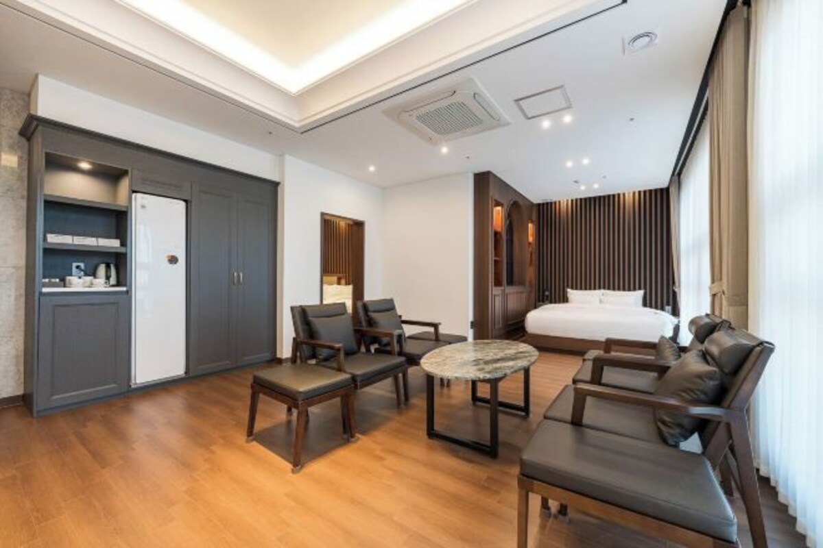 Una habitación de hotel moderna con una cama, un rincón de estar con sofás y una mesa de centro, una kitchenette y un ventanal grande.